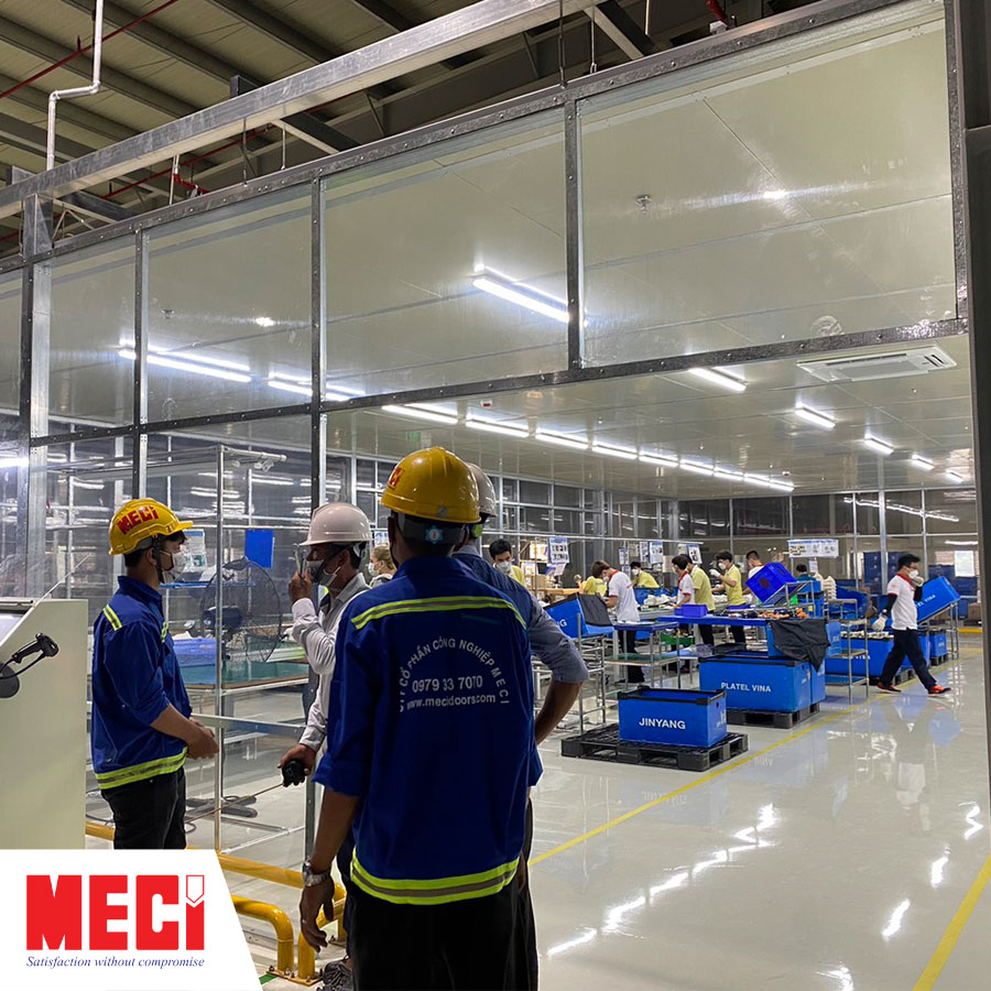 4 anh nhân viên meci đang thảo luận phương án trước một xưởng phân loại sợi, bên trong có nhiều nam nhân viên đang làm việc