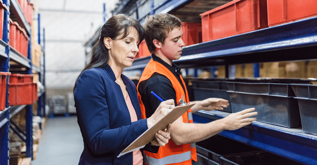 Trong một nhà xưởng, người đàn ông đang kiểm tra thùng hàng, người phụ nữ đang đứng bên cạnh ghi chép vô hồ sơ