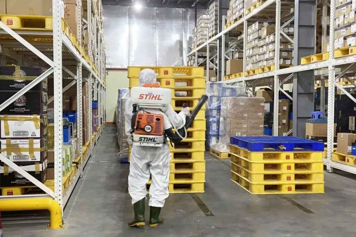 một người mặc đồ bảo hộ đang đeo bình thuốc diệt côn trùng và cầm vòi xịt đi giữa kho hàng
